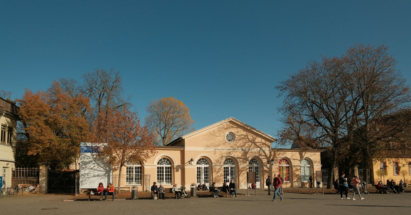 Menschen schlendern über einen sonnigen Platz vor einem historischen Gebäude.